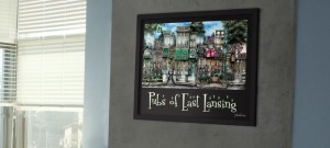A framed artwork titled "pubs of east lansing" depicting various bars, displayed on a wall between two windows with blinds.
